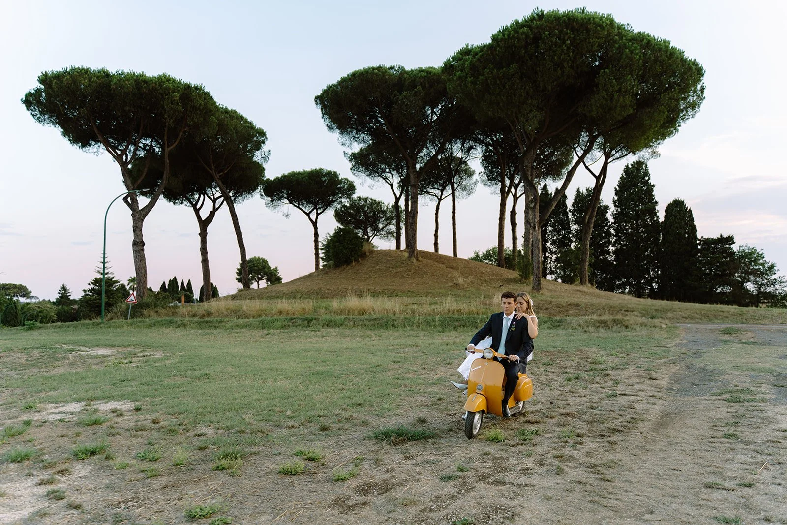 A couple in formal attire riding a yellow scooter on a dirt path in a park with tall trees in the background.