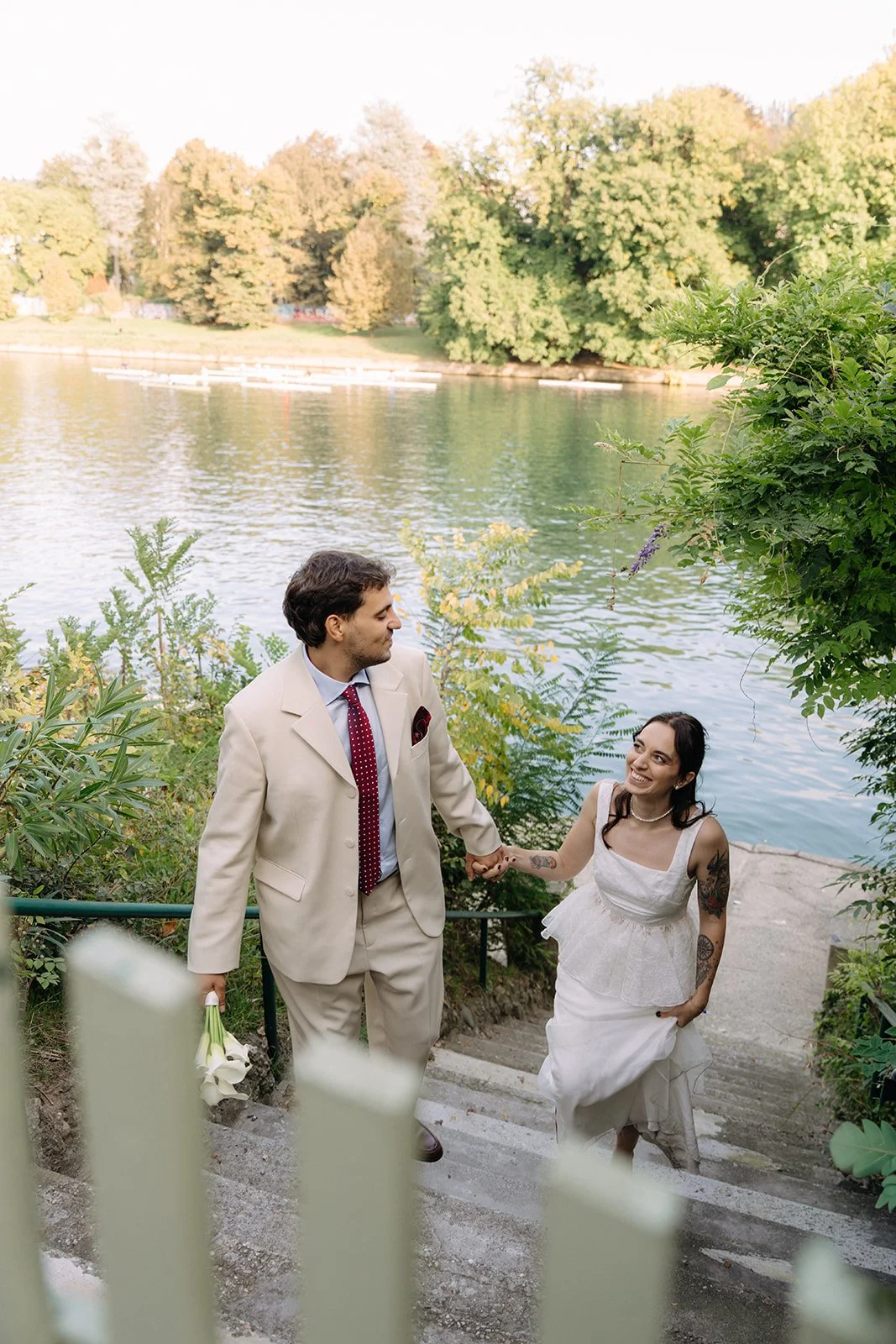 A couple, dressed in wedding attire, holding hands and ascending a staircase near a body of water with trees in the background.
