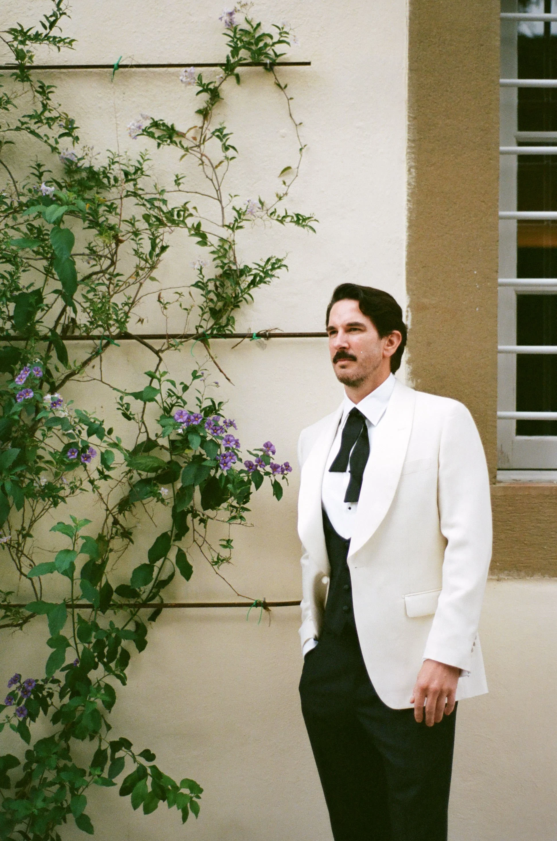 A man in a white tuxedo jacket, black vest, and black tie standing outdoors near a beige wall with greenery, purple flowers, and a window with metal bars.