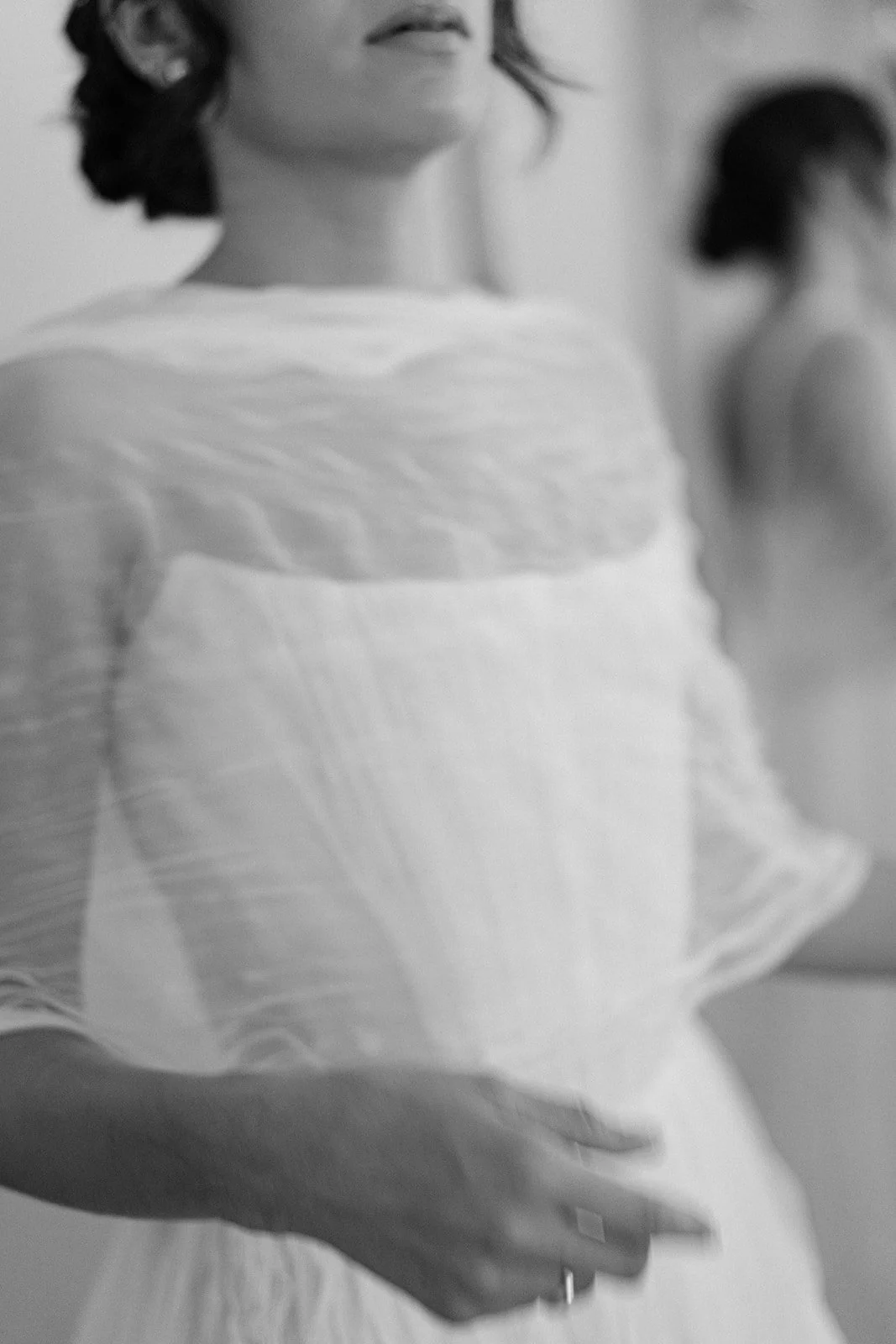 A woman with short dark hair and a ring on her left hand, wearing a light-colored, textured top, standing with her hand on her hip. In the background, a blurred woman with hair tied back.