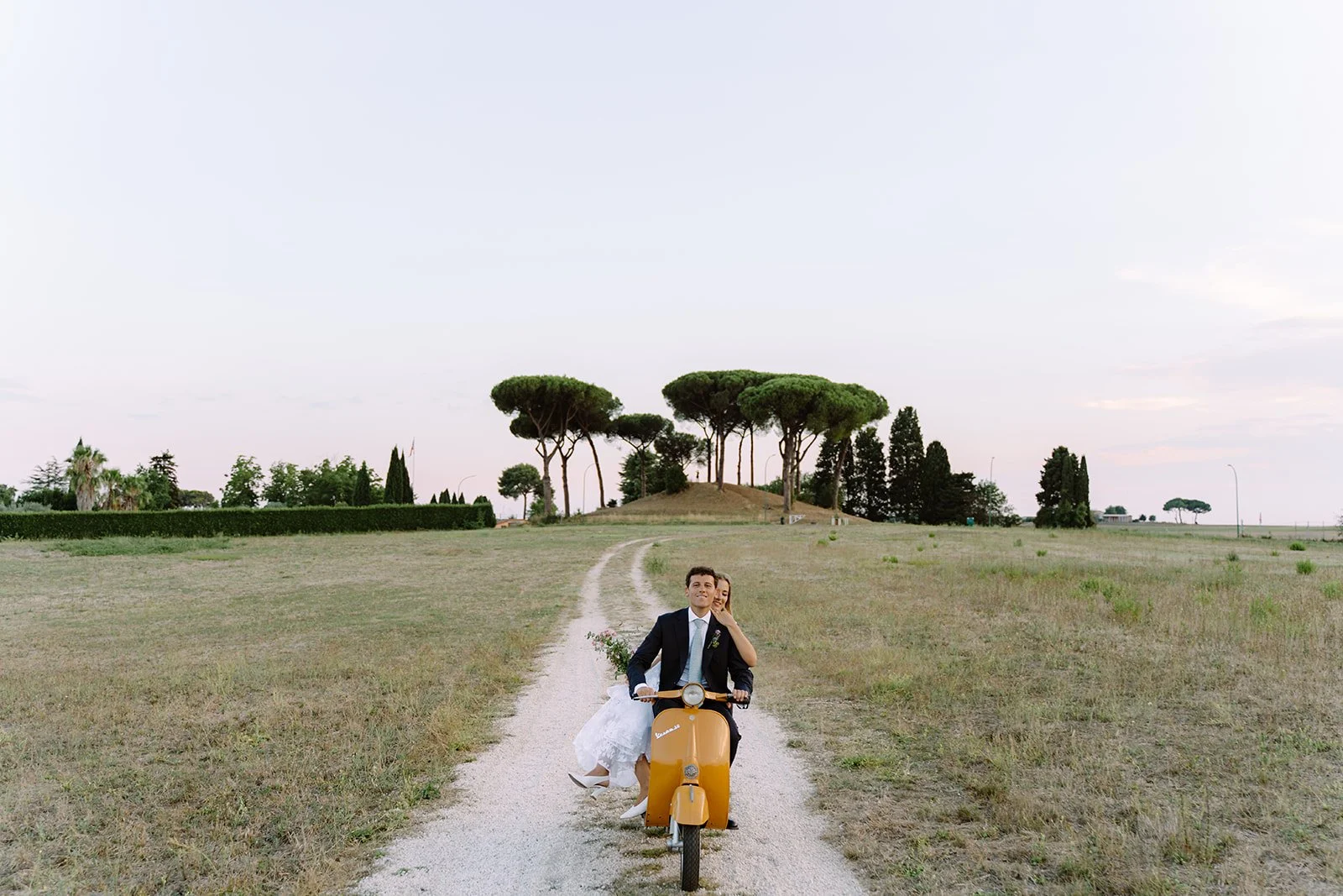 A newlywed couple riding a yellow scooter on a dirt path in a rural landscape with trees and open sky.