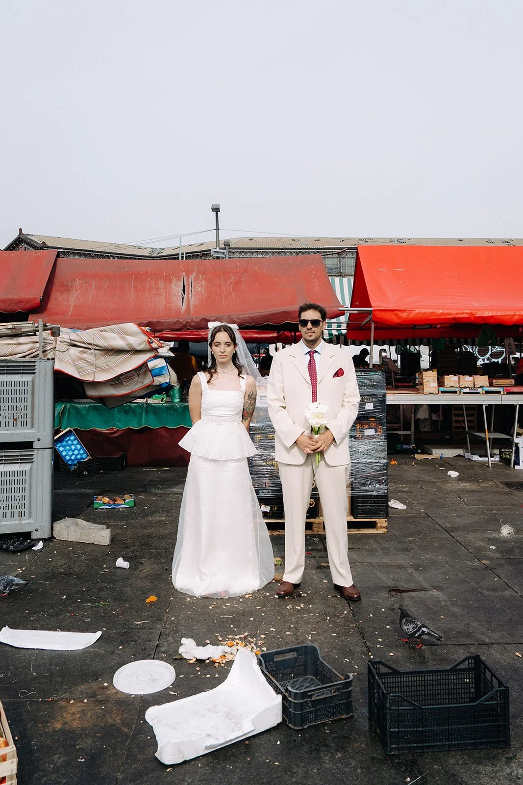A man and woman dressed in wedding attire standing in front of a market stall with red awnings. The woman is wearing a white wedding dress and veil, and the man is in a cream suit with a red tie, holding white flowers. The ground is littered with tra