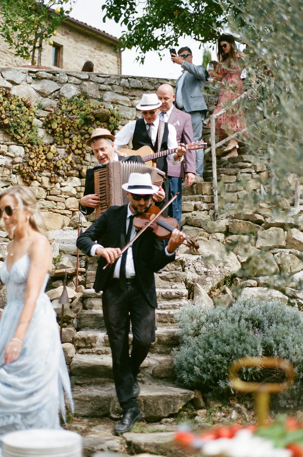 A group of musicians playing instruments descending stone steps during an outdoor gathering, with guests standing on the steps listening.