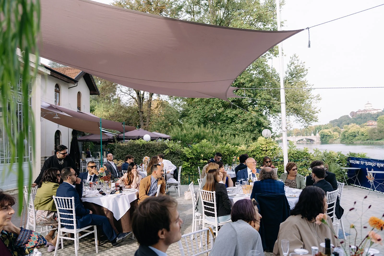 People gathered at outdoor event on patio with tables, chairs, and shade sails, near river with bridge and building on hillside in background.