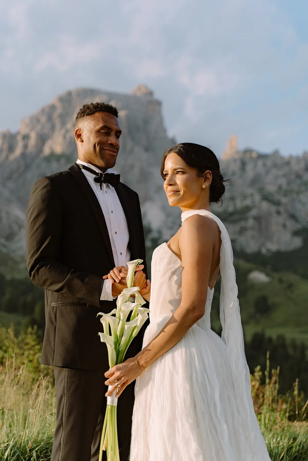 A bride and groom holding hands outdoors, with mountains in the background during sunset. The bride wears a white wedding dress and holds a bouquet of white calla lilies; the groom wears a black tuxedo.