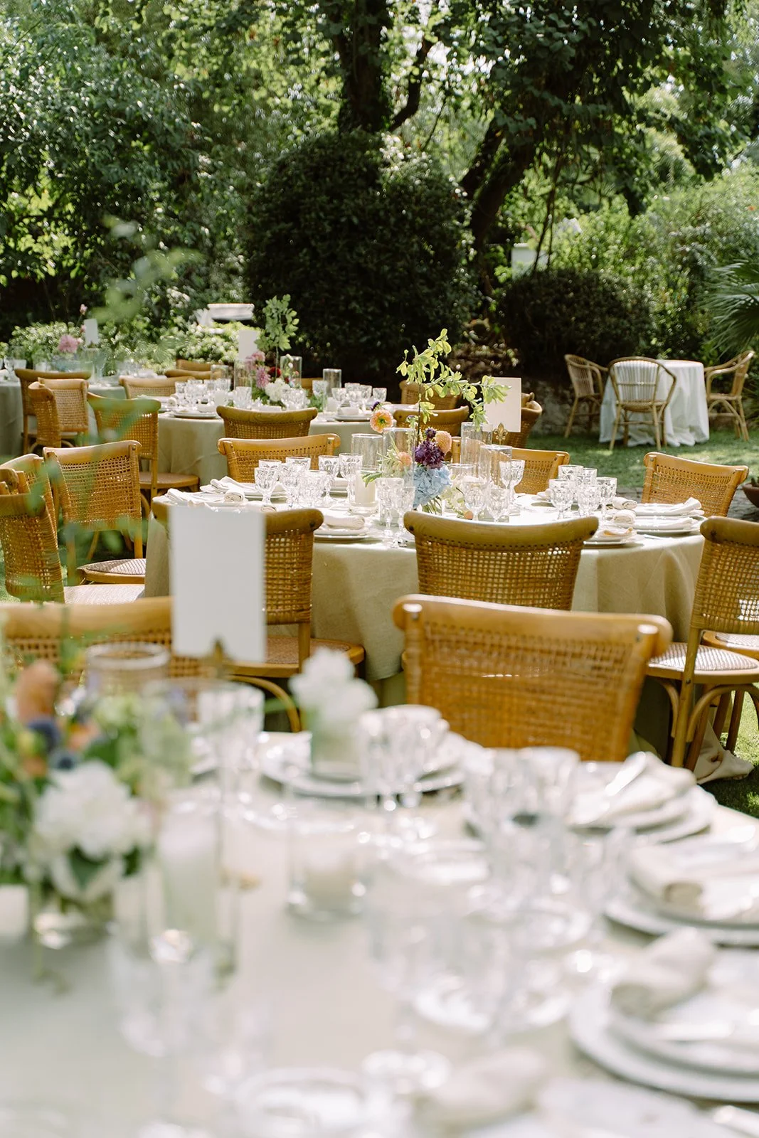 Outdoor wedding reception setup with round tables covered in cream tablecloths, decorated with floral centerpieces and surrounded by wicker chairs on a lush green garden.