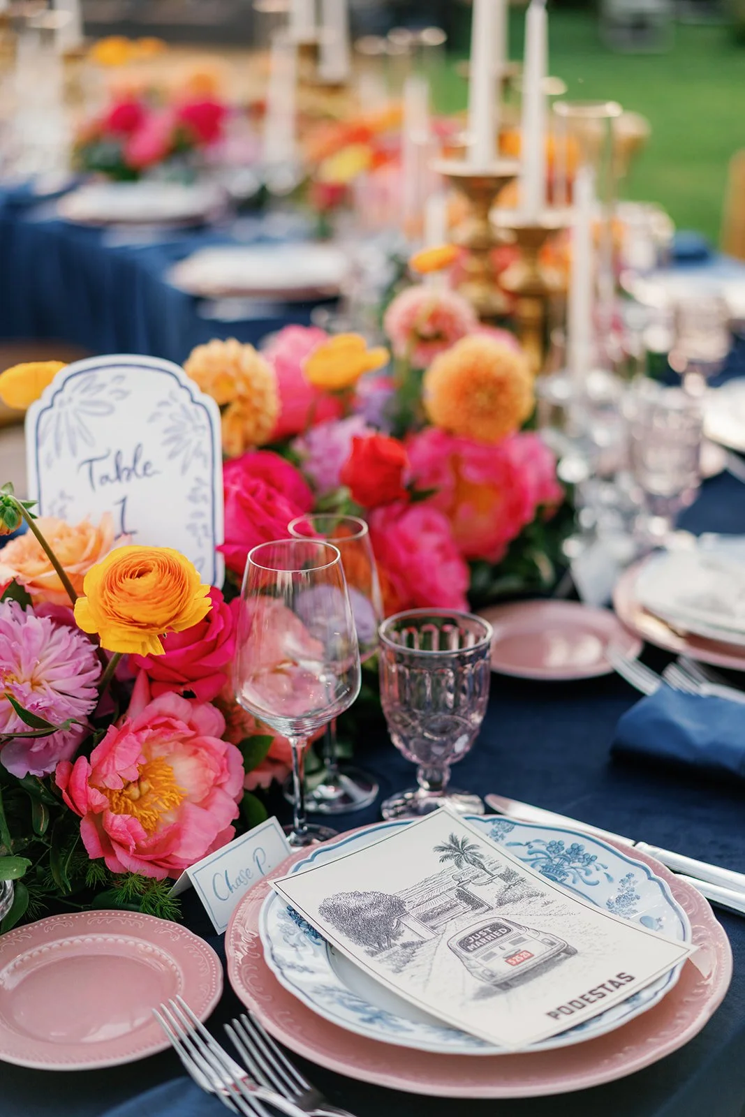 A beautifully decorated outdoor dining table with pink, yellow, and peach flowers, pink and clear glassware, pink plates, and a navy tablecloth. There are place cards with handwritten names and a sign indicating 'Table 1'.