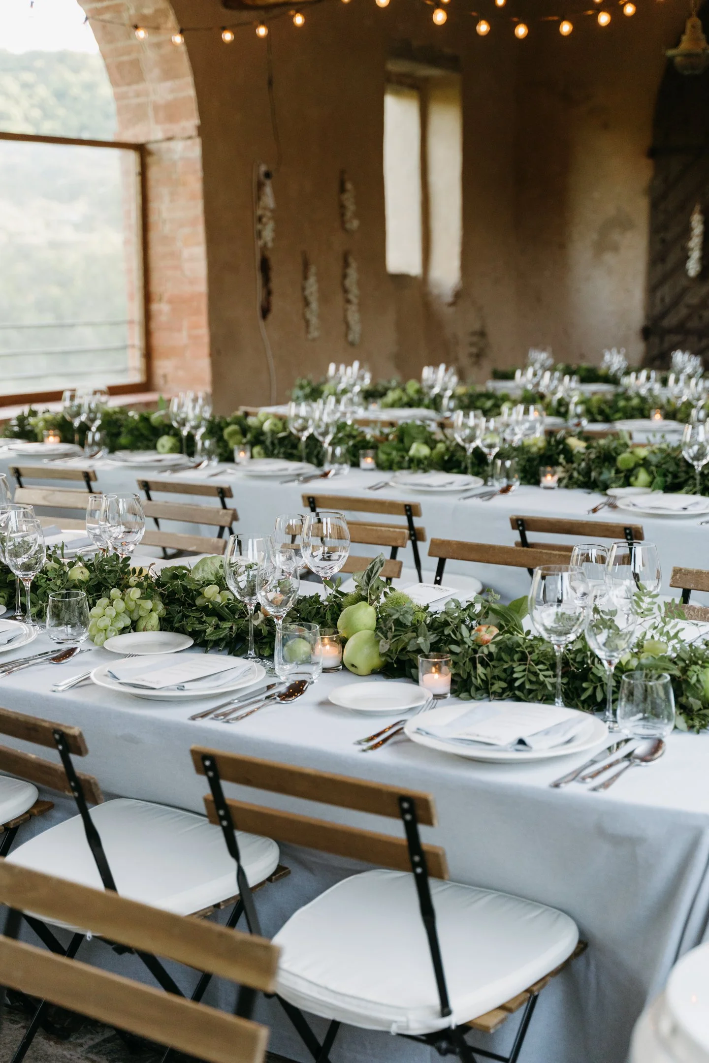 A beautifully set banquet table with white linens, decorated with greenery, apples, grapes, candles, wine glasses, and white plates, arranged for an event in a rustic venue with exposed brick and string lights.