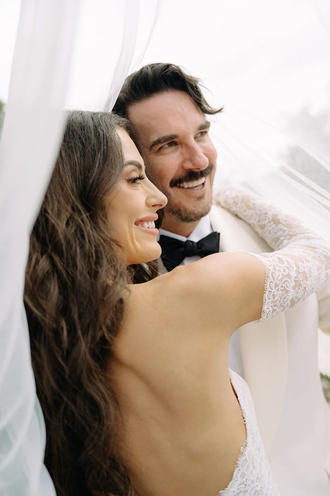 A happy bride and groom smiling close together on their wedding day, with the bride's arm around the groom's shoulder.