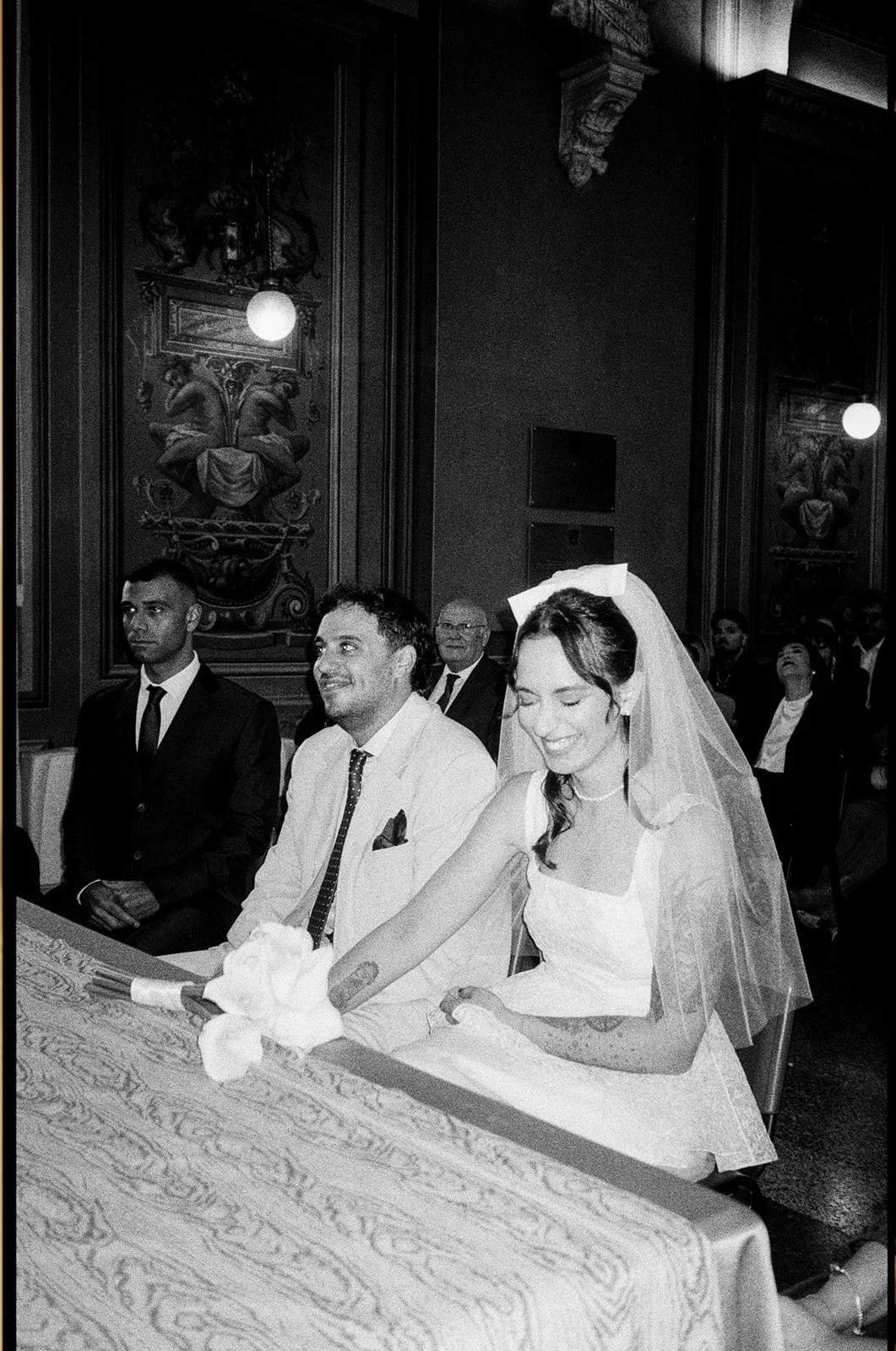 A black and white photo of a wedding ceremony with the bride and groom sitting at a table, smiling. The bride is wearing a veil and a white dress, and the groom is in a light-colored suit. Several guests are seated in the background.