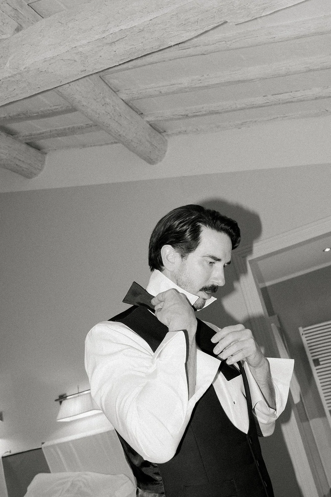 A man with dark hair and a mustache adjusts his collar while wearing a tuxedo with a white shirt, in a room with modern decor.