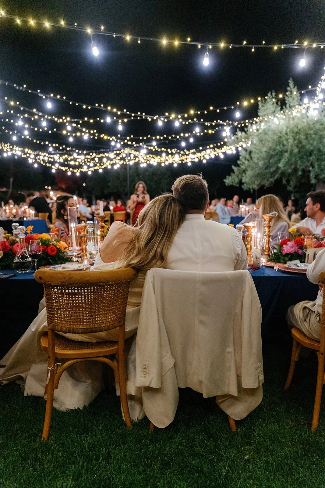 Couple sitting closely together at an outdoor nighttime dinner party under string lights, with multiple tables decorated with flowers and candles, surrounded by other guests.