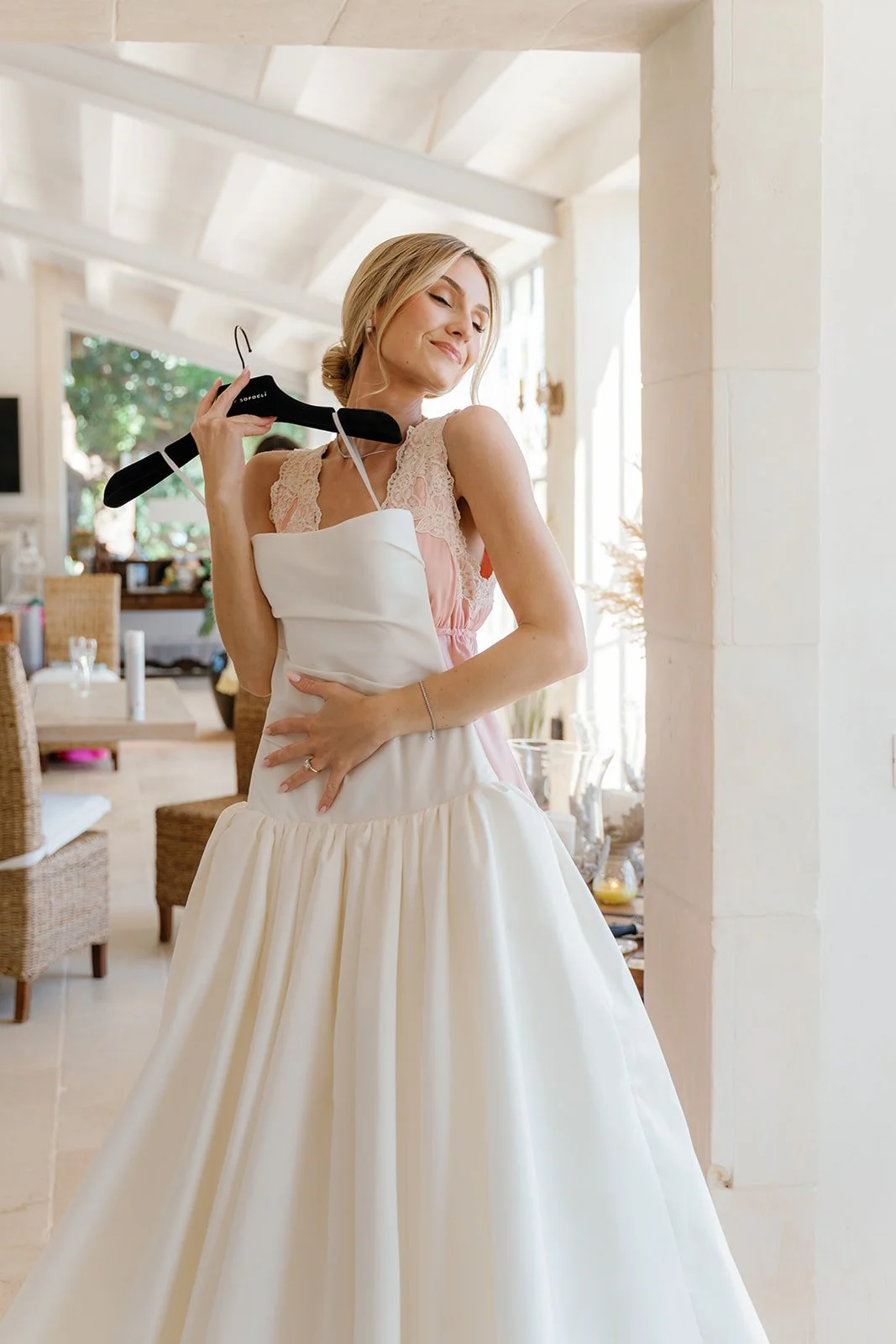 A woman in a wedding dress happily hugging a wedding gown hanger inside a bright, decorated room.