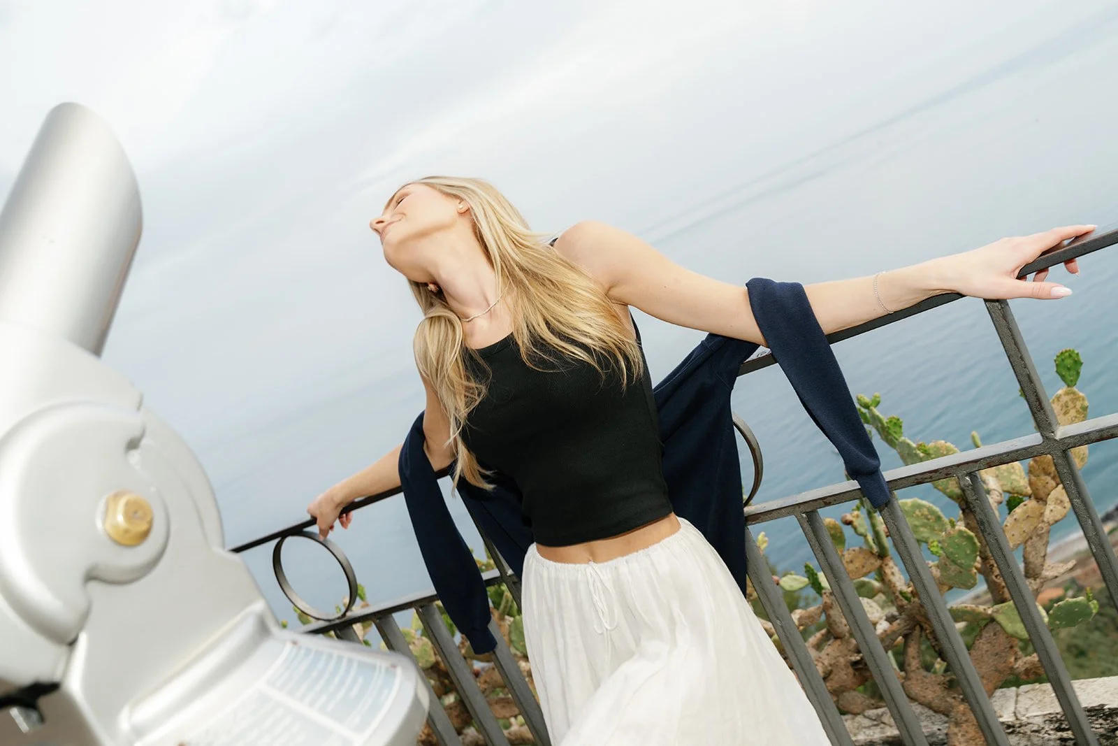 A woman with long blonde hair leaning against a metal railing near the water, with cactus plants behind her and a coin-operated viewing telescope in the foreground.