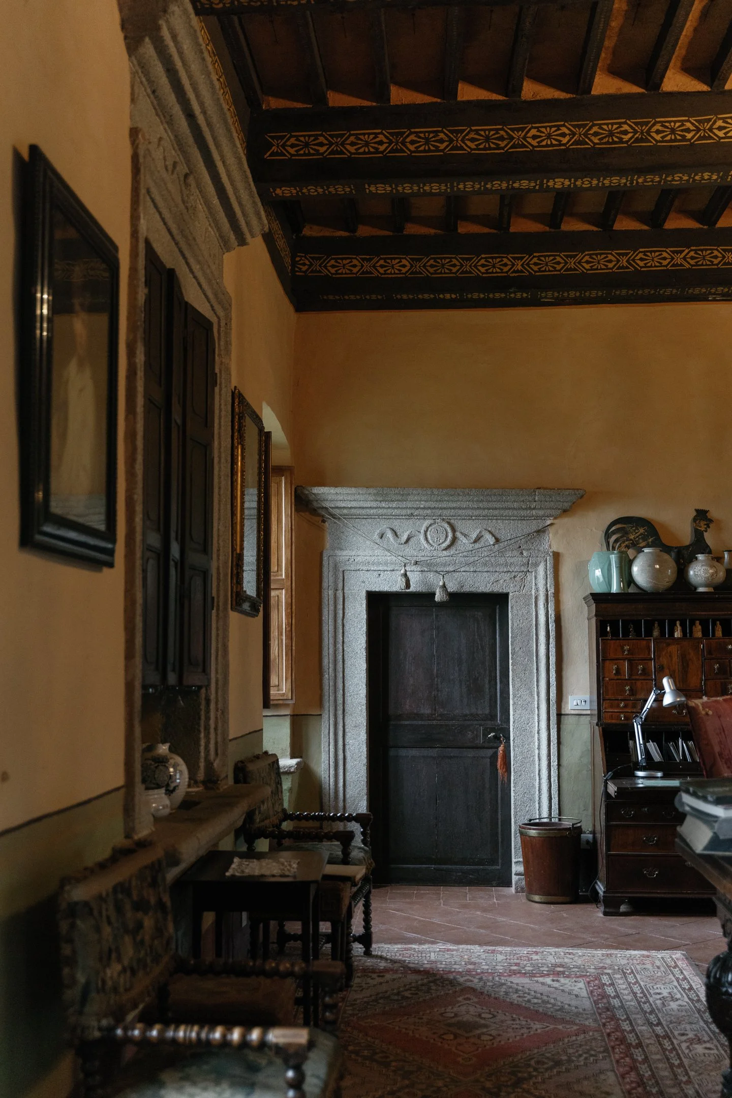 Interior of an antique room with a stone doorframe, wooden ceiling beams with decorative patterns, vintage chairs, a dark wooden cabinet with vases, a rug, and warm colored walls.