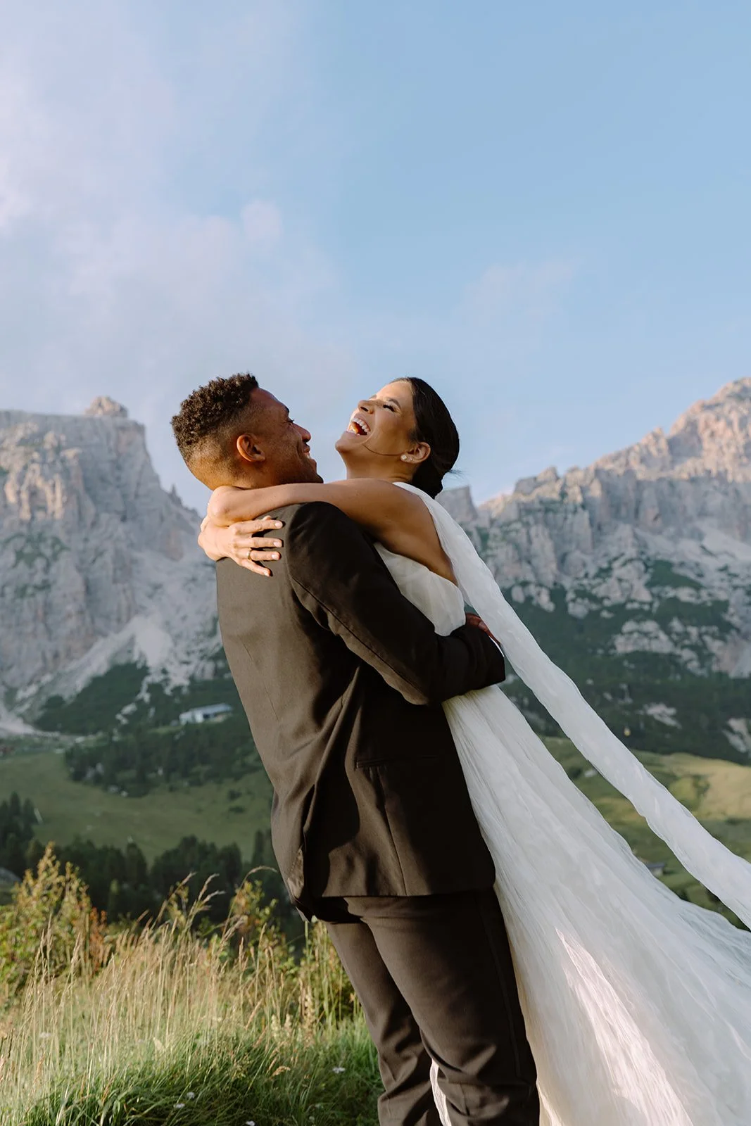 A couple dressed in wedding attire embracing outdoors with mountains and blue sky in the background.