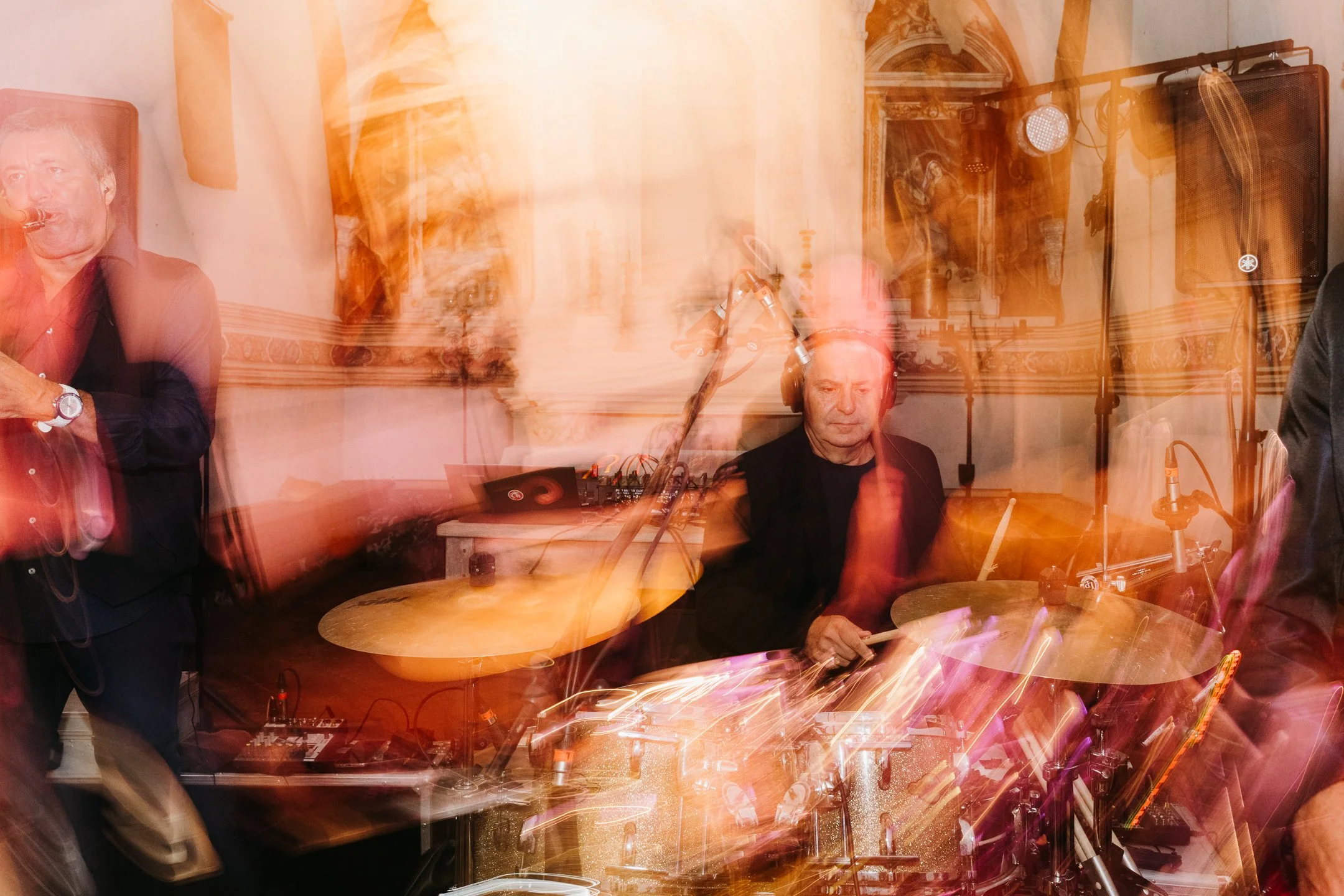 A man playing drums at a live music event in an ornate room with gold accents and artwork.