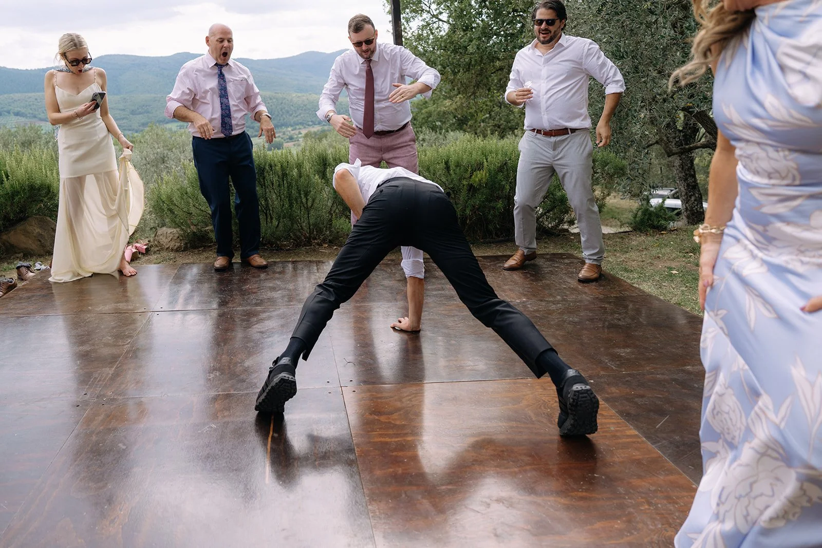 A man in a white shirt and black pants is breakdancing on a wooden dance floor outdoors, surrounded by five people watching and reacting enthusiastically.