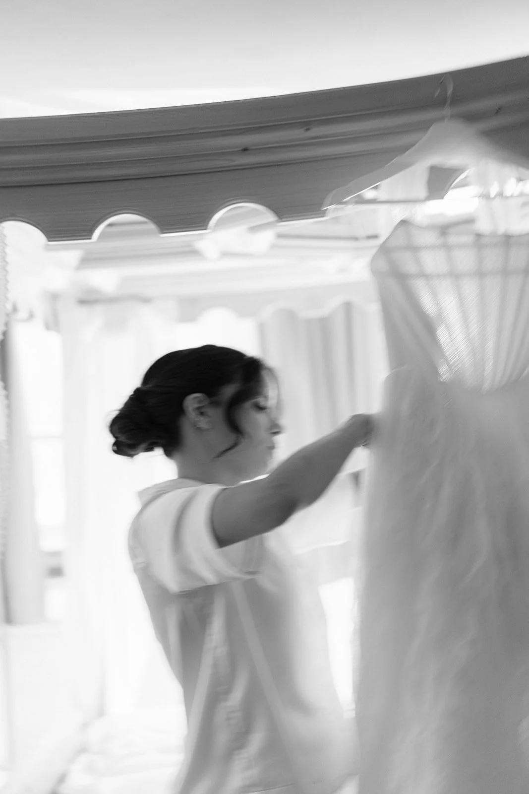 A woman with dark hair styled in an updo, wearing a light-colored shirt, is folding or hanging a large piece of fabric or clothing indoors.