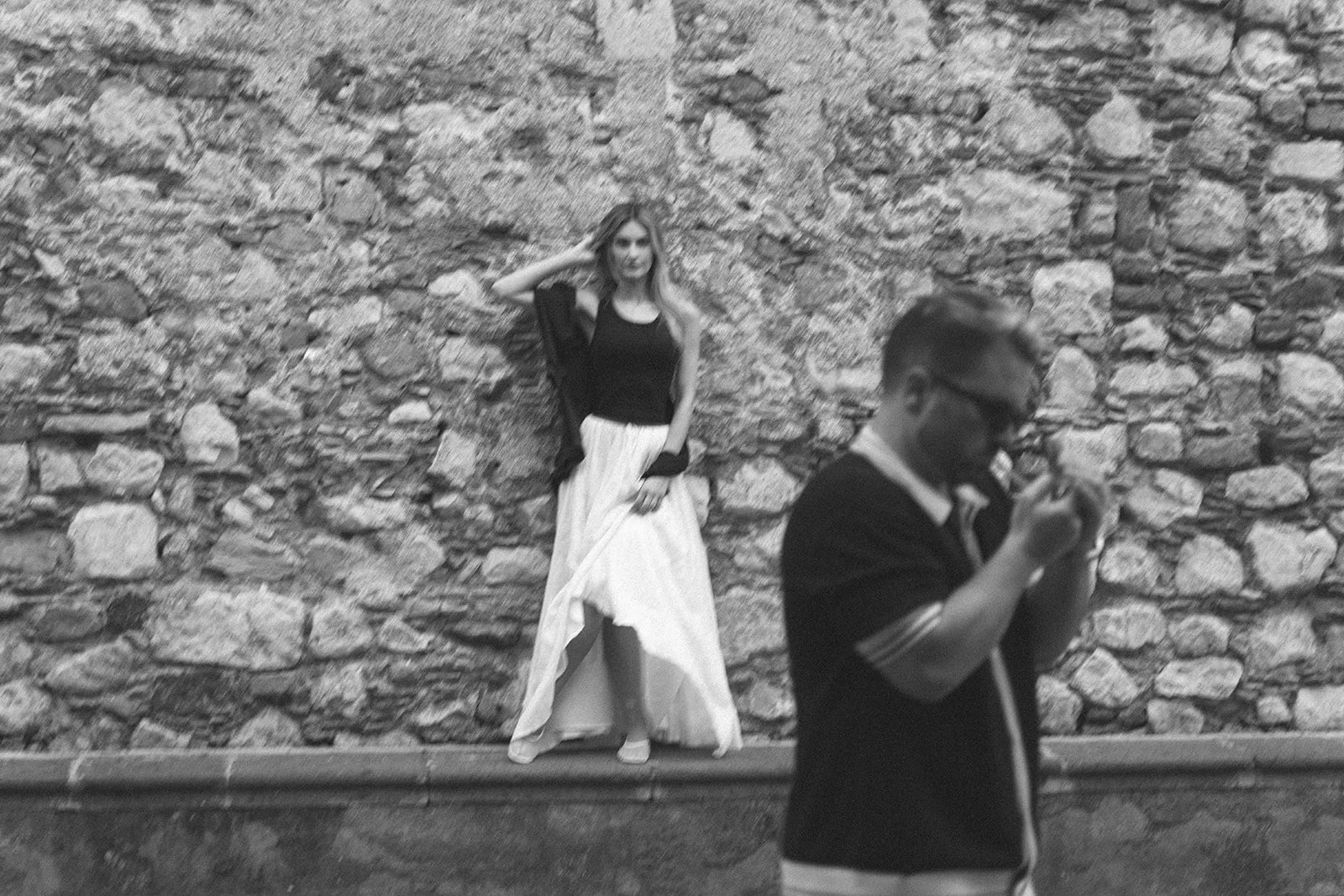 A woman in a black top and long white skirt leaning against a stone wall, with a man in a dark shirt and glasses in the foreground looking at his phone.