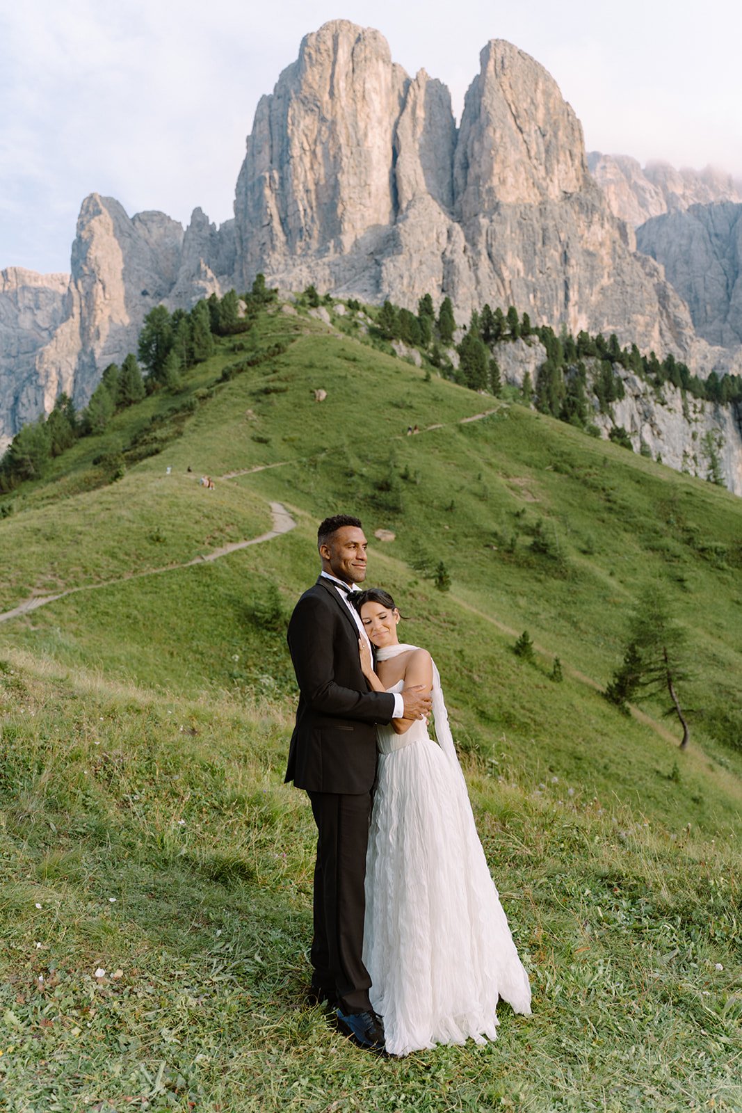 A newlywed couple in wedding attire standing on a grassy hillside with a large mountain and green hill in the background.