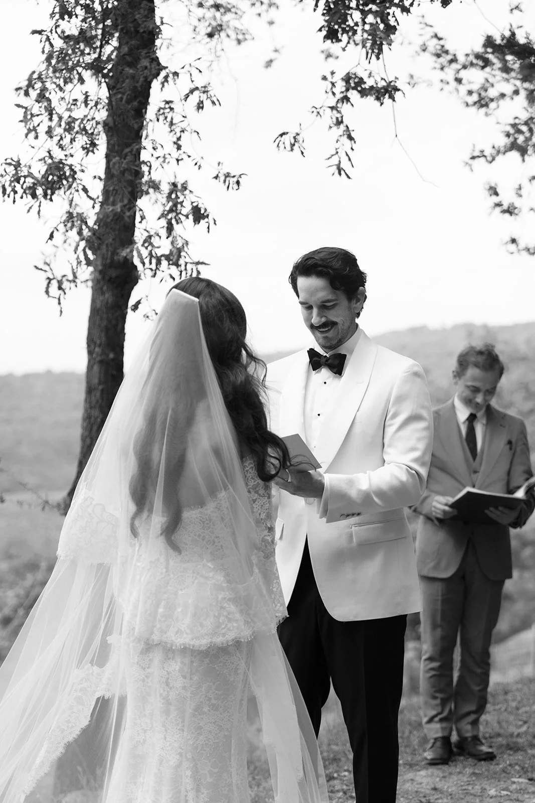 A black-and-white photo of a wedding ceremony outdoors, with a bride and groom exchanging vows under a tree, and a man in a suit reading from a book in the background.