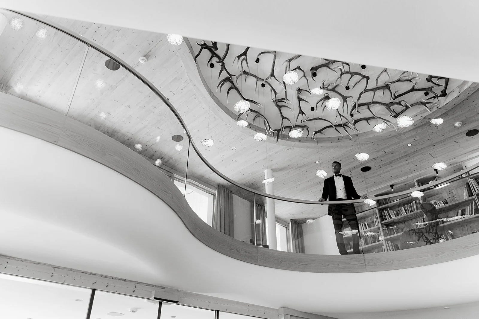 A man in a tuxedo standing on a balcony in a modern, stylish building with a decorative ceiling, chandeliers, and a bookshelf.