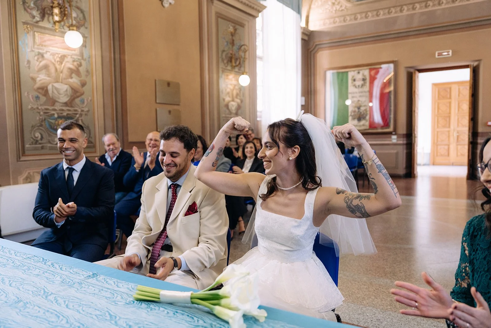 A woman in a wedding dress and veil, with tattoos, flexing her arms and smiling, seated at a table with a bouquet of white calla lilies. She is inside a grand, ornately decorated room with an audience clapping and smiling in the background.