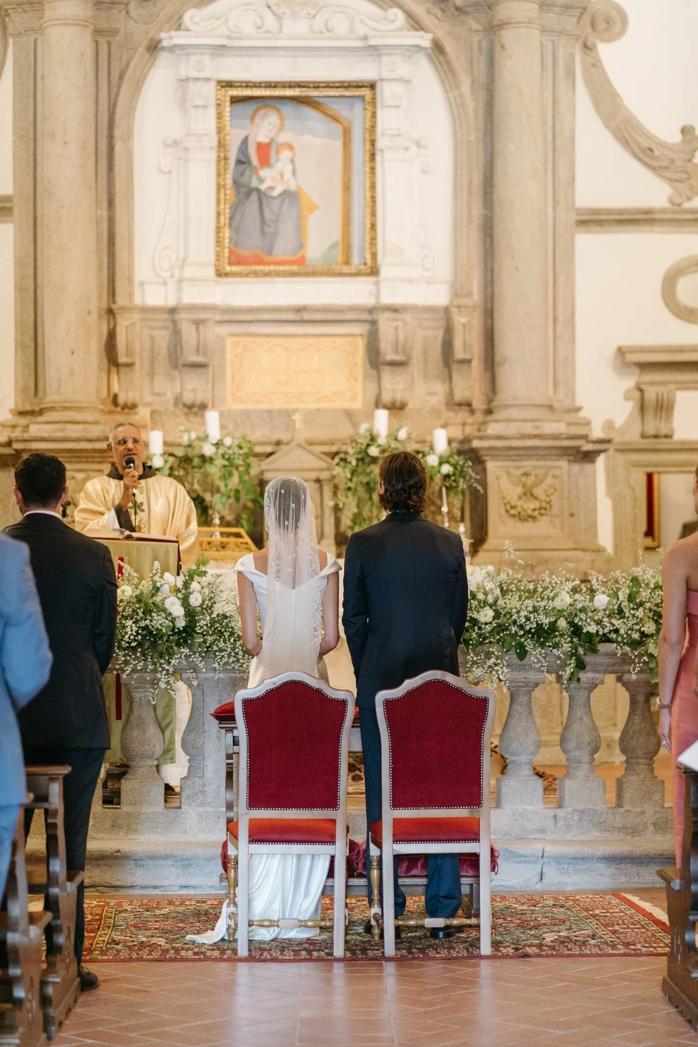 A wedding ceremony inside a church, with the bride and groom standing at the altar facing a priest, during the vows. A painting of Madonna and Child is hung above the altar decorated with white flowers and candles. Guests are seated, and some are sta