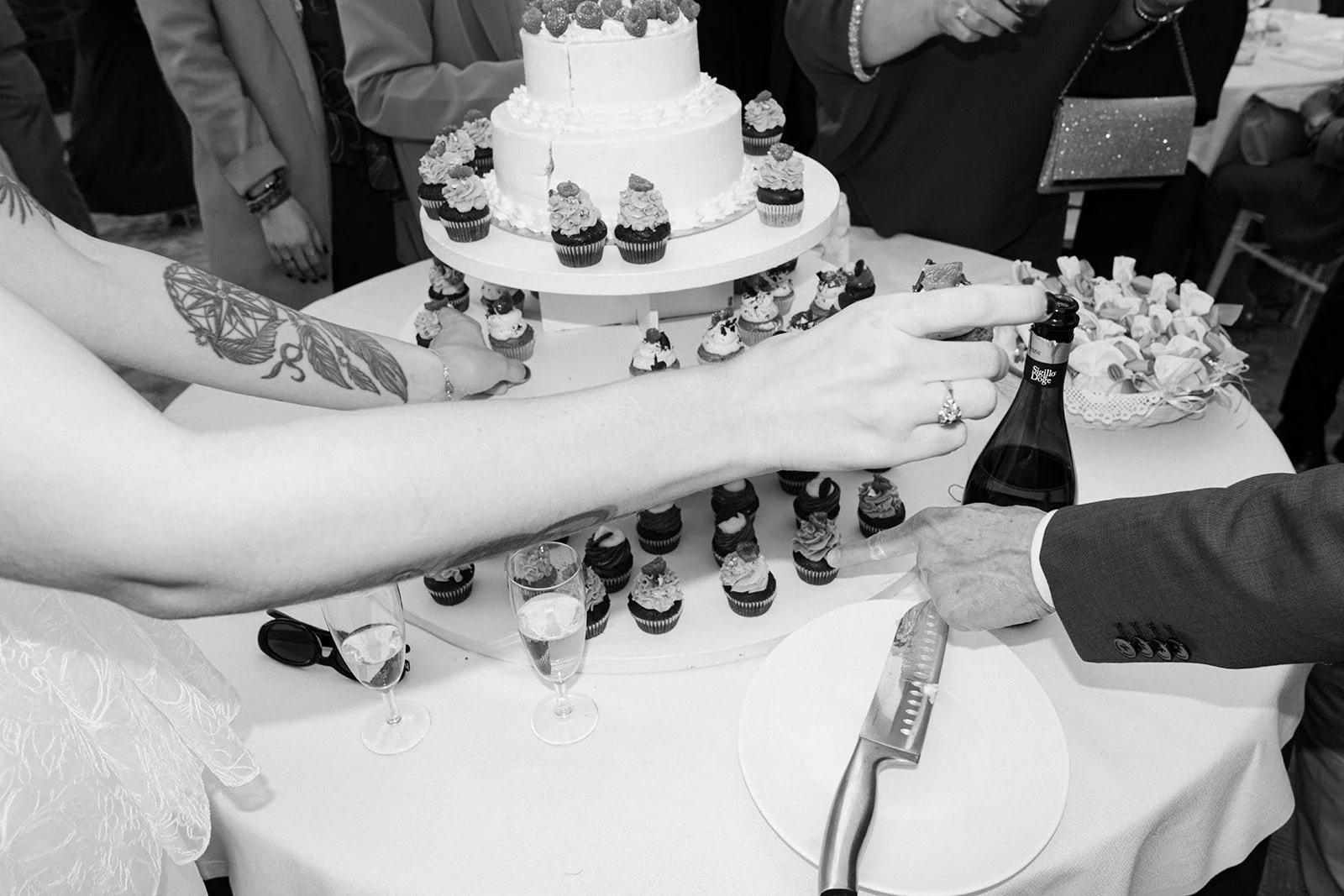 A wedding cake and numerous cupcakes on a tiered stand on a table. Two people are cutting the cake together, one holding a knife and the other holding a bottle of champagne. Champagne glasses are on the table, and guests are visible in the background