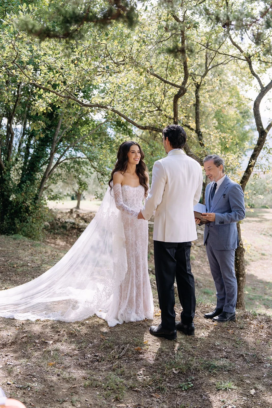 A bride and groom holding hands during a wedding ceremony outdoors, with an officiant standing beside them, under a tree in a natural setting.