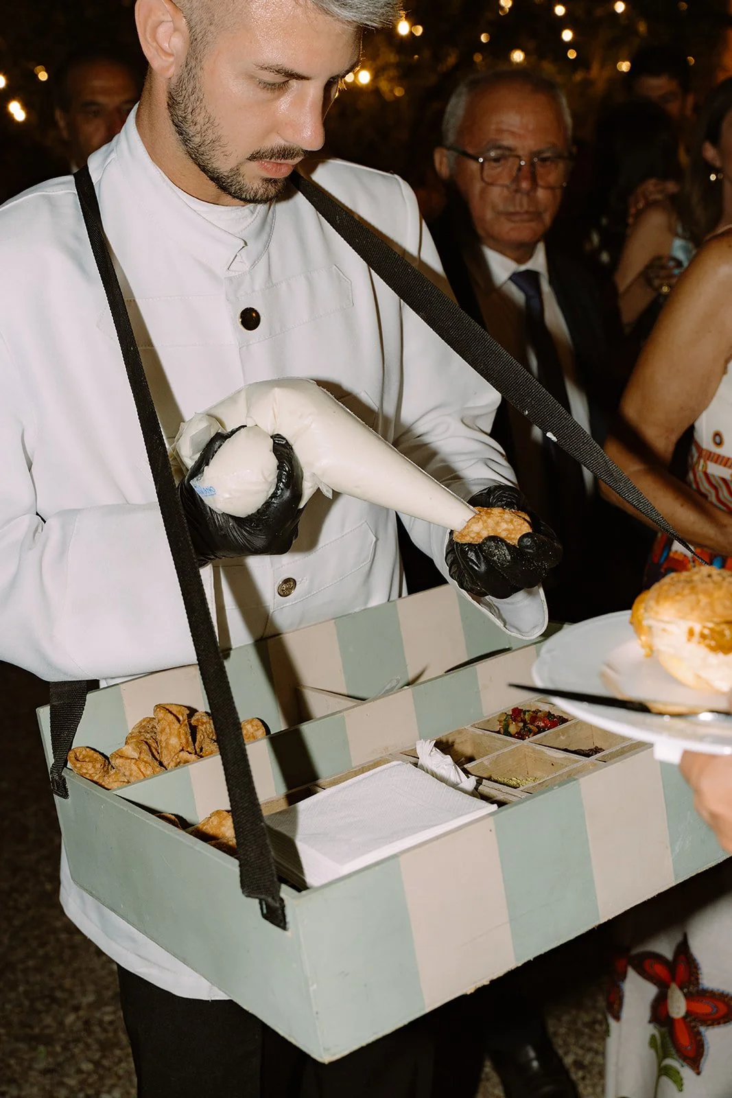 A chef wearing a white coat and black gloves is filling a pastry bag with cream. The chef is attending a dessert station with a tray of pastries and toppings on display, and several people are in the background at what appears to be a party or event.