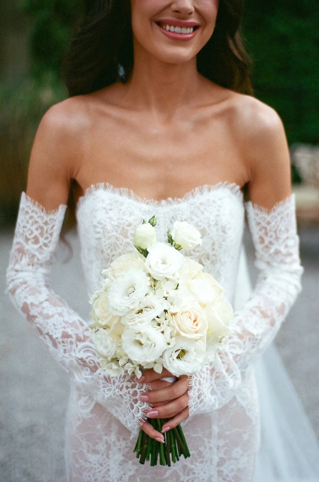 A woman in a white lace wedding dress holding a bouquet of white and cream-colored roses.