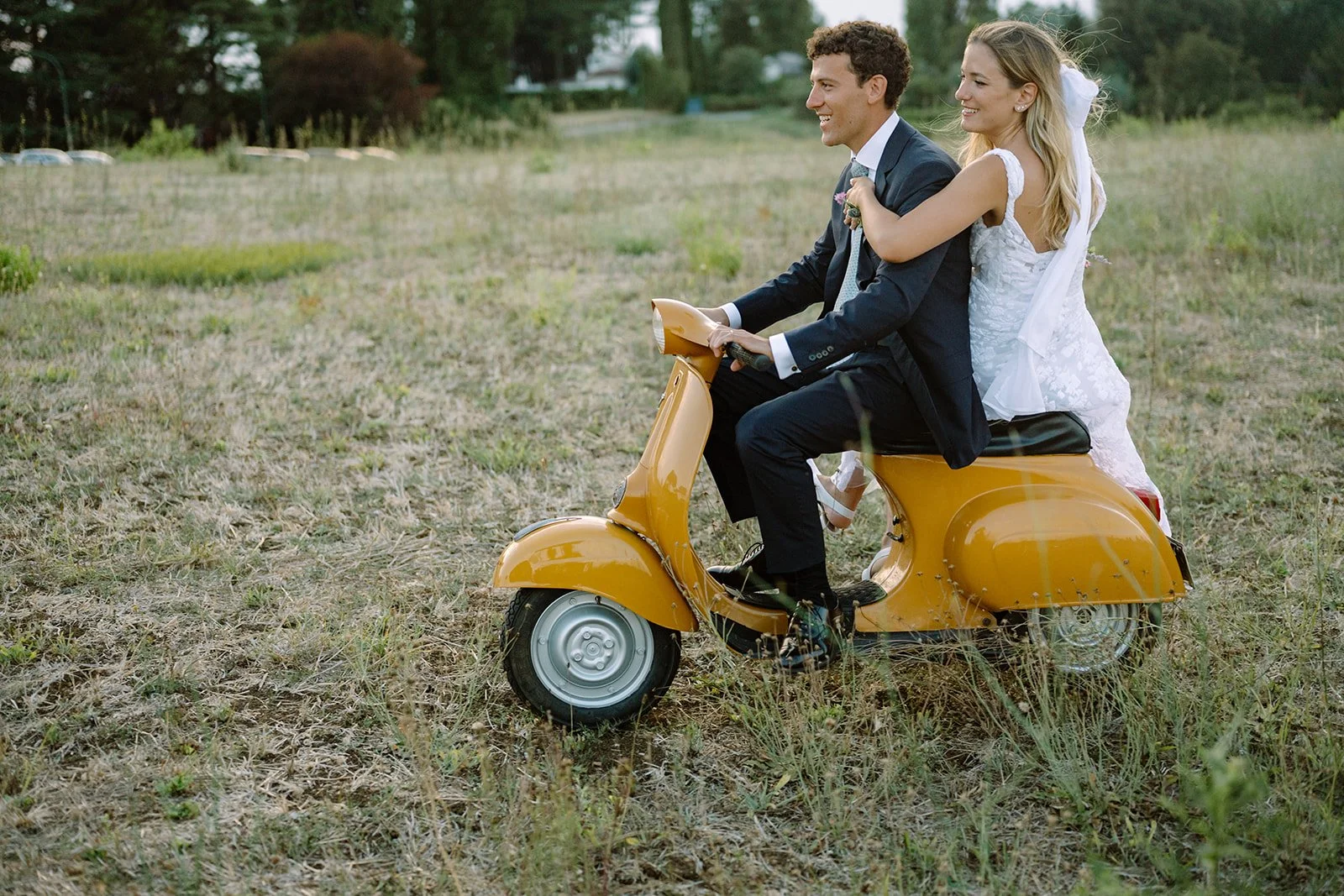 A bride and groom in wedding attire riding on a yellow scooter in an open grassy field.