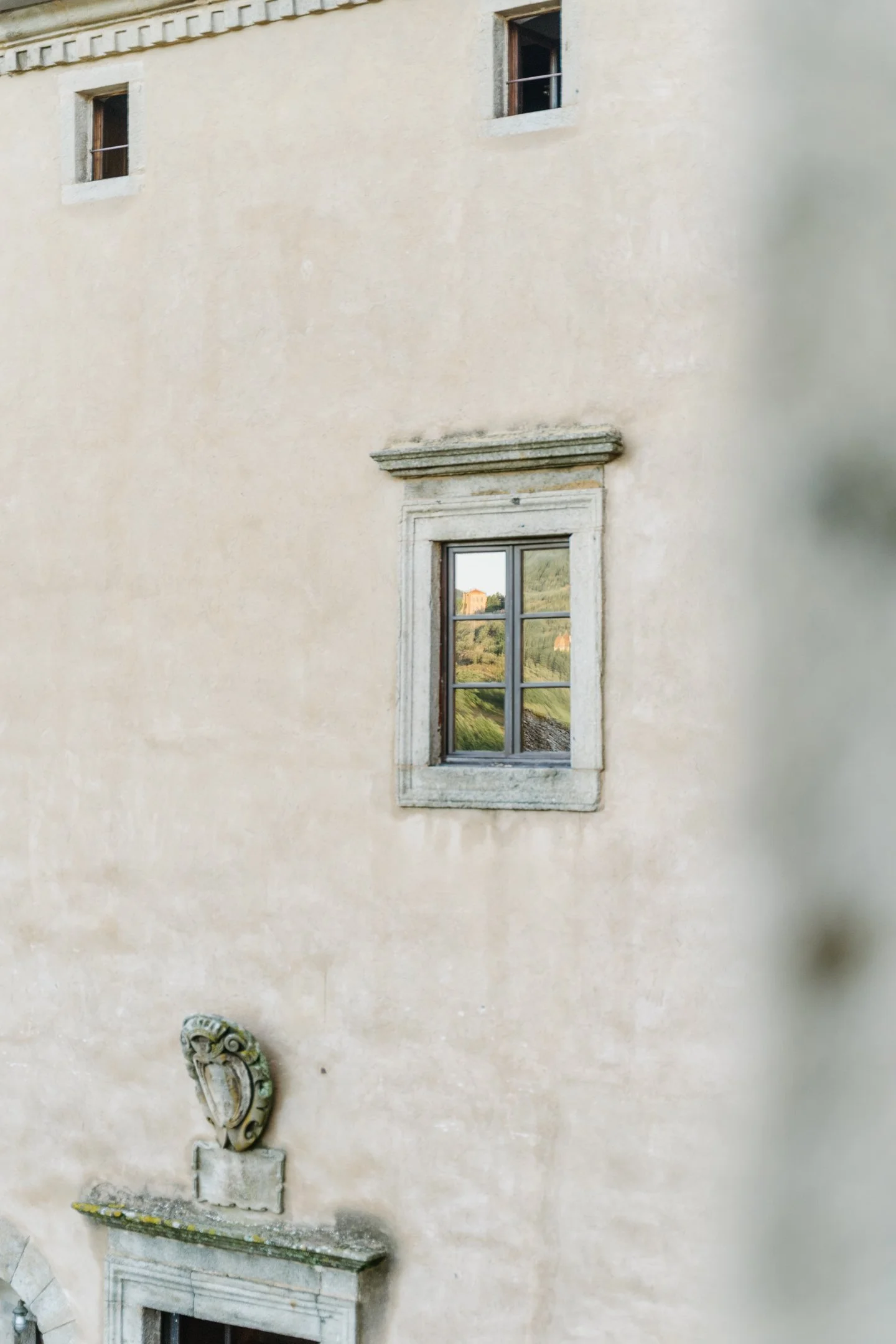 A beige stone wall of a historic building with two small upper windows, a window with a view of greenery and hills, and a decorative stone shield above a door.