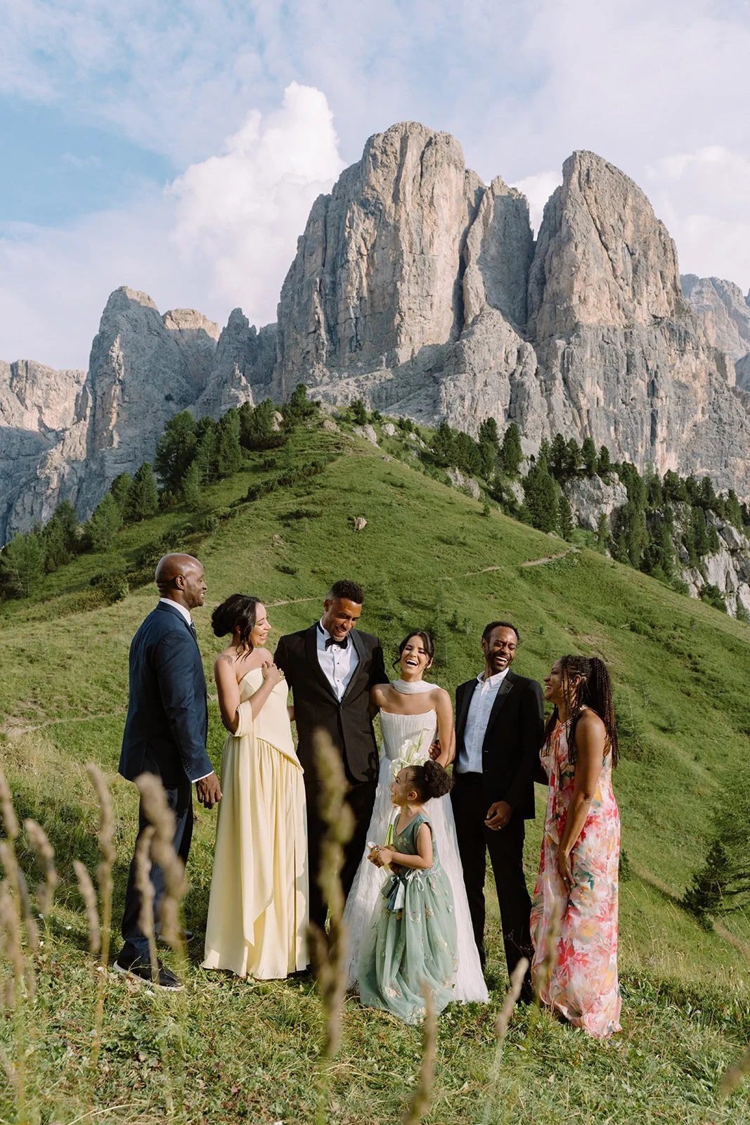 A group of seven people, including two children, standing together outdoors on a grassy hillside with a mountainous landscape in the background. They are dressed in semi-formal and casual attire, and appear to be laughing and enjoying each other's co