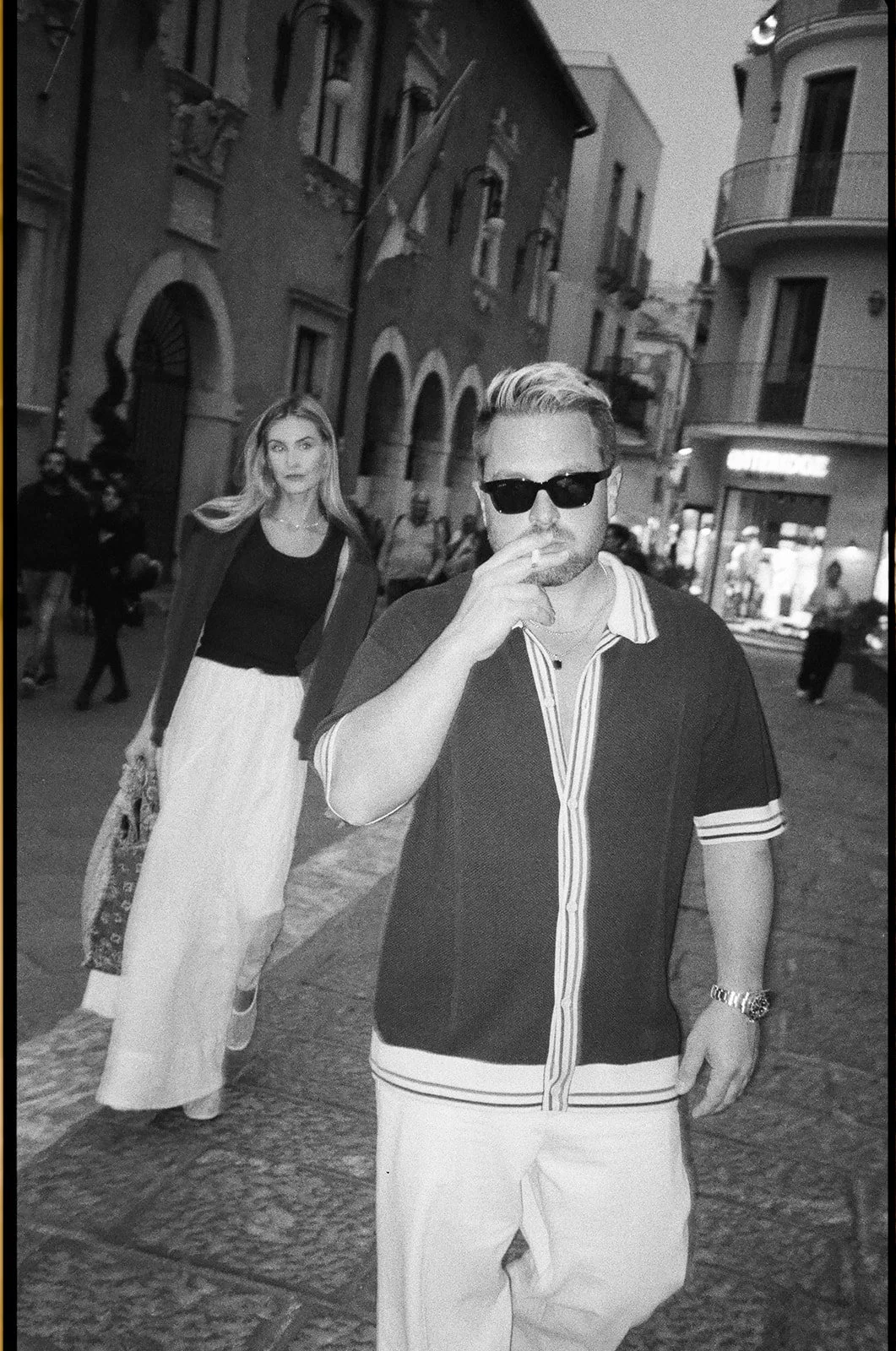 A black and white photo of a man wearing sunglasses, a striped shirt, and light-colored pants, standing on a city street, smoking a cigarette, with a woman walking behind him. The woman has long hair, a dark top, and a long skirt, carrying a bag, and