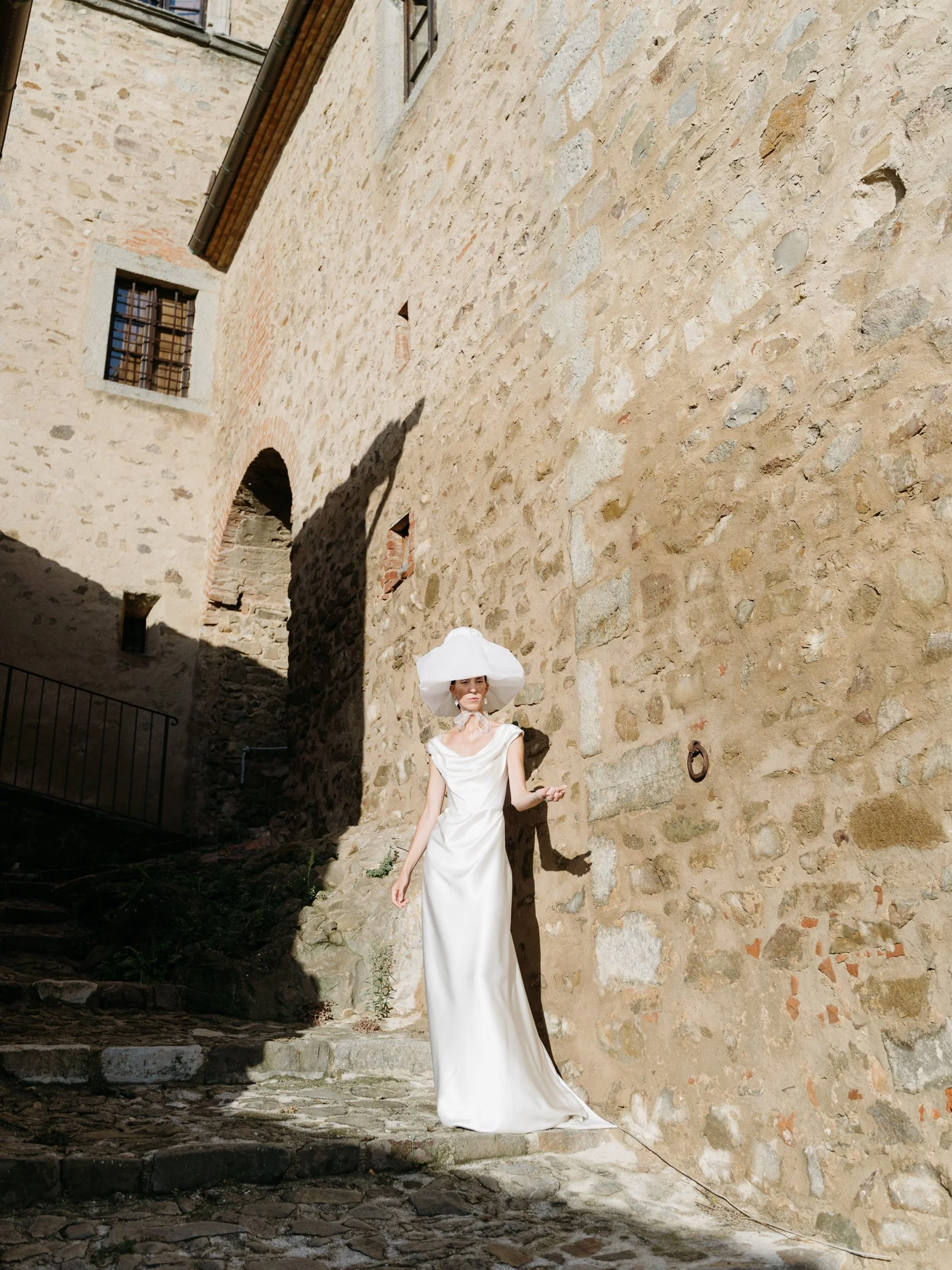 A woman dressed in a white gown with a wide-brimmed hat, standing on a cobblestone street against a stone wall with small windows, casting shadows in a sunny outdoor setting.