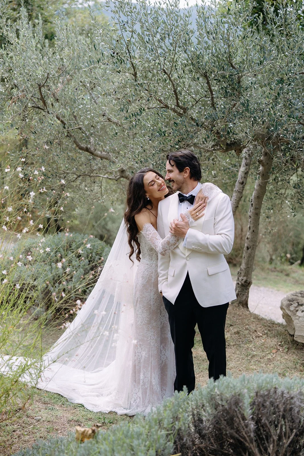 A bride and groom share a joyful moment outdoors among trees and greenery, with the bride wearing a lace wedding dress and veil, and the groom in a white tuxedo jacket and black bow tie.