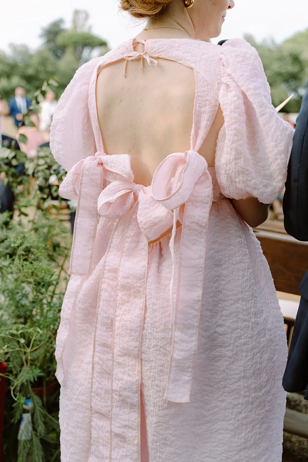 Close-up of a woman wearing a light pink dress with puffed sleeves, open back, and tied bows, at an outdoor event with trees and people in the background.