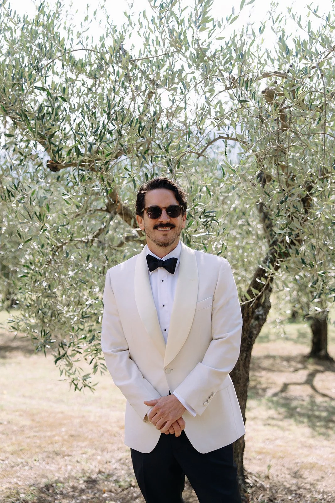 A man dressed in a white tuxedo jacket, black bow tie, and sunglasses stands outdoors in front of a tree with green leaves, smiling.