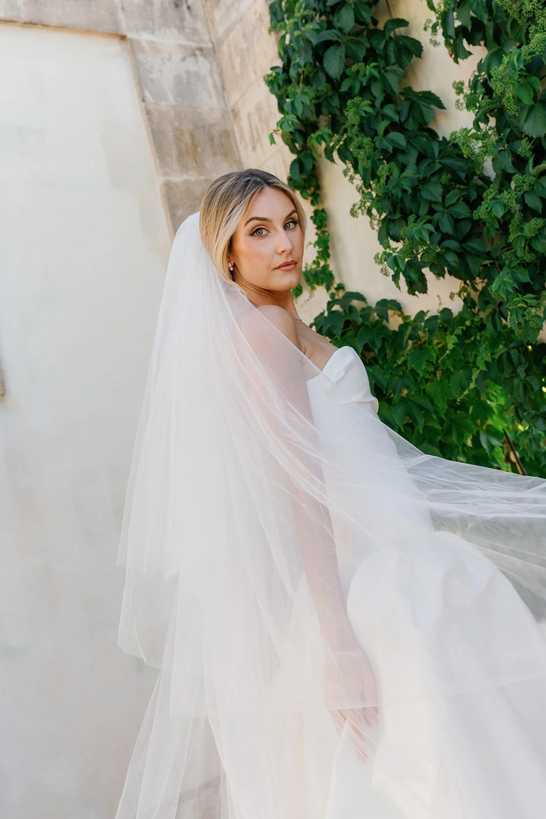 Bridal portrait of a woman in a white wedding gown with a veil, standing outdoors next to green foliage.