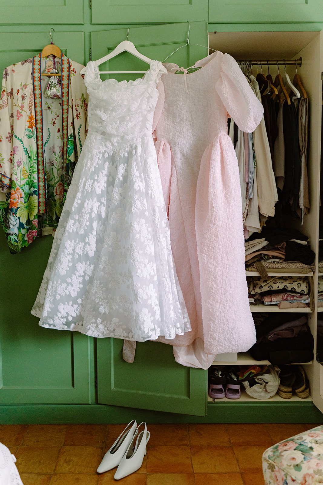 A closet with various dresses and clothing items hanging, including a white lace dress, a light pink dress, a floral patterned dress, and other garments. A pair of white high heels is on the floor in front of the closet.
