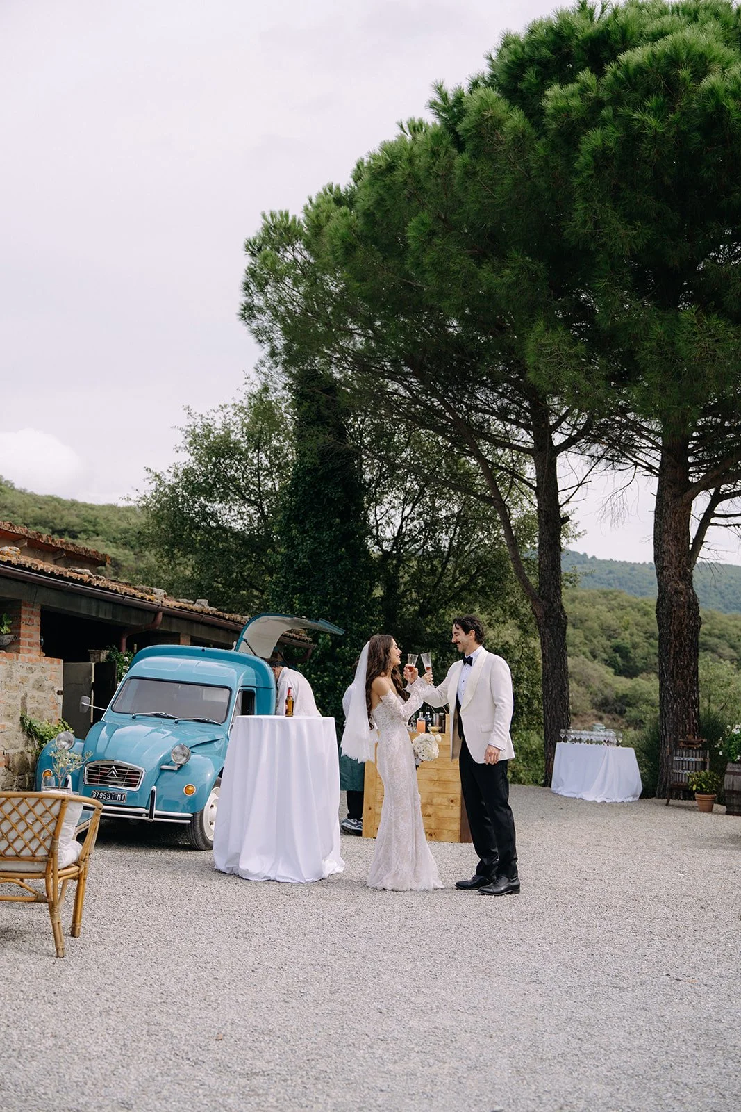 A bride and groom celebrating with drinks at their wedding reception outdoors, with a vintage blue car and scenic greenery in the background.