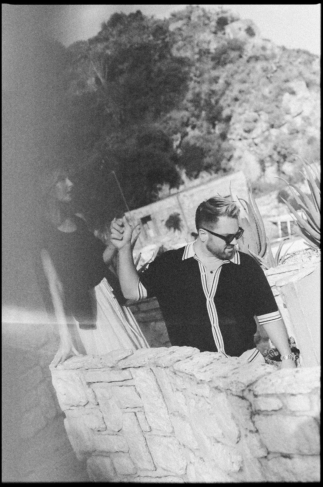 A black and white photo of a man with sunglasses and a trimmed beard, wearing a striped button-up shirt, holding a thin branch in one hand and resting his other hand on a stone wall. Behind him, a woman with long dark hair is leaning on the wall. In 