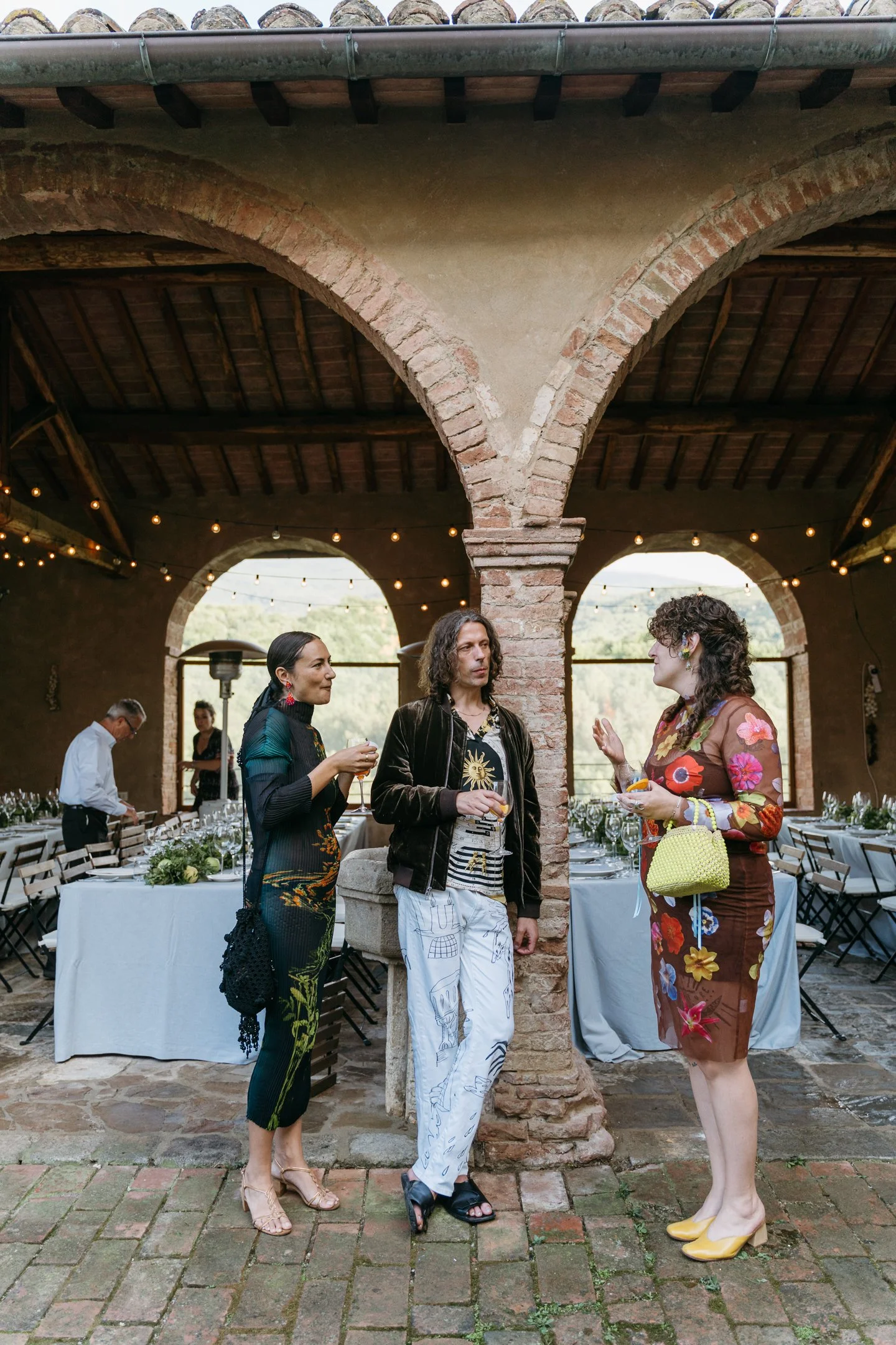 Three people are standing and talking in an outdoor covered dining area decorated with string lights. The woman on the left is wearing a black dress with colorful patterns and nude heels. The man in the middle has curly hair and is dressed in a black