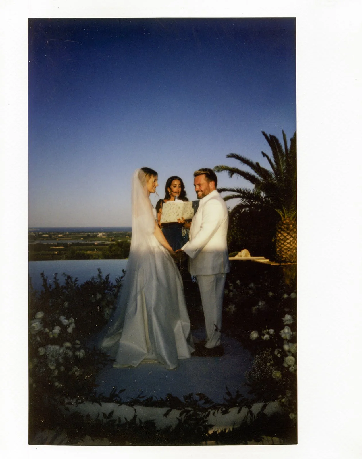 A couple getting married outdoors during the evening with a bride in a white wedding dress and veil, a groom in a white suit, and a woman officiating, with palm trees and a body of water in the background.