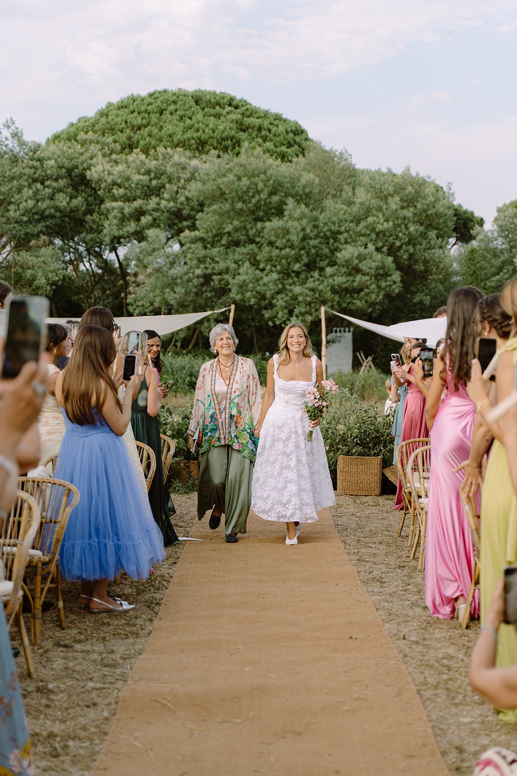 A bride walking down an outdoor wedding aisle, holding a bouquet of pink flowers, surrounded by friends and family taking photos. The scene is set in a lush green garden with a large tree in the background.
