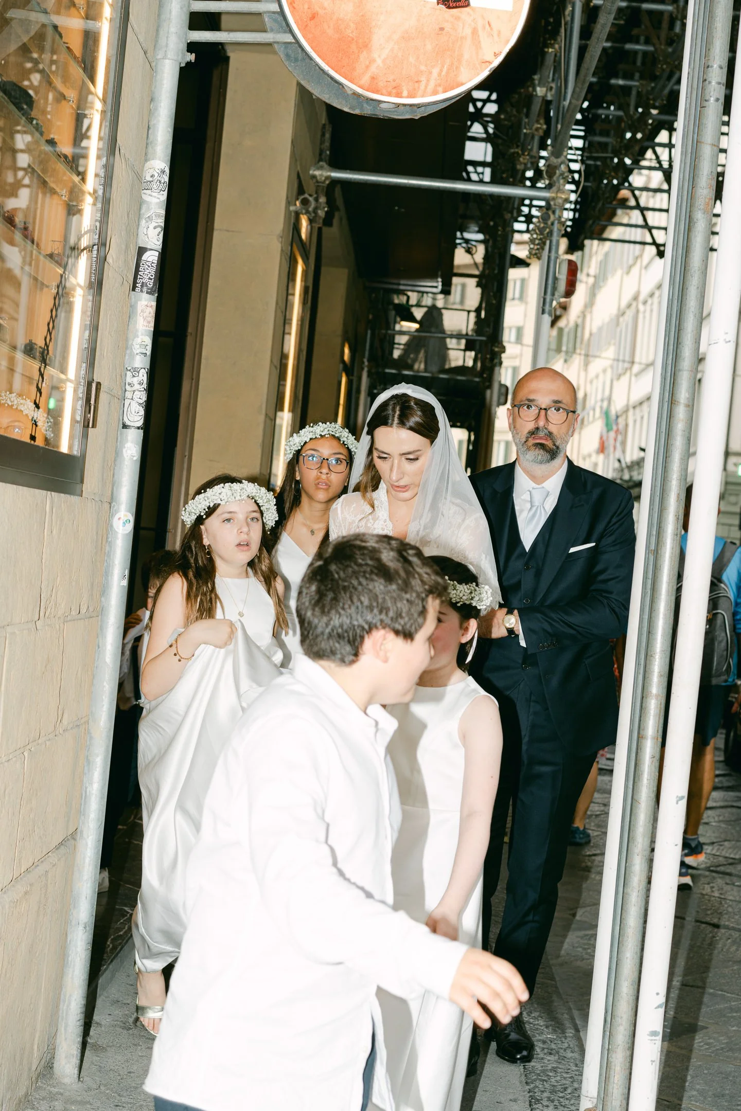 A wedding procession walking on a city sidewalk, featuring a bride in a white wedding gown and veil, children in white clothes and flower crowns, and a man in a dark suit with a white shirt and tie.