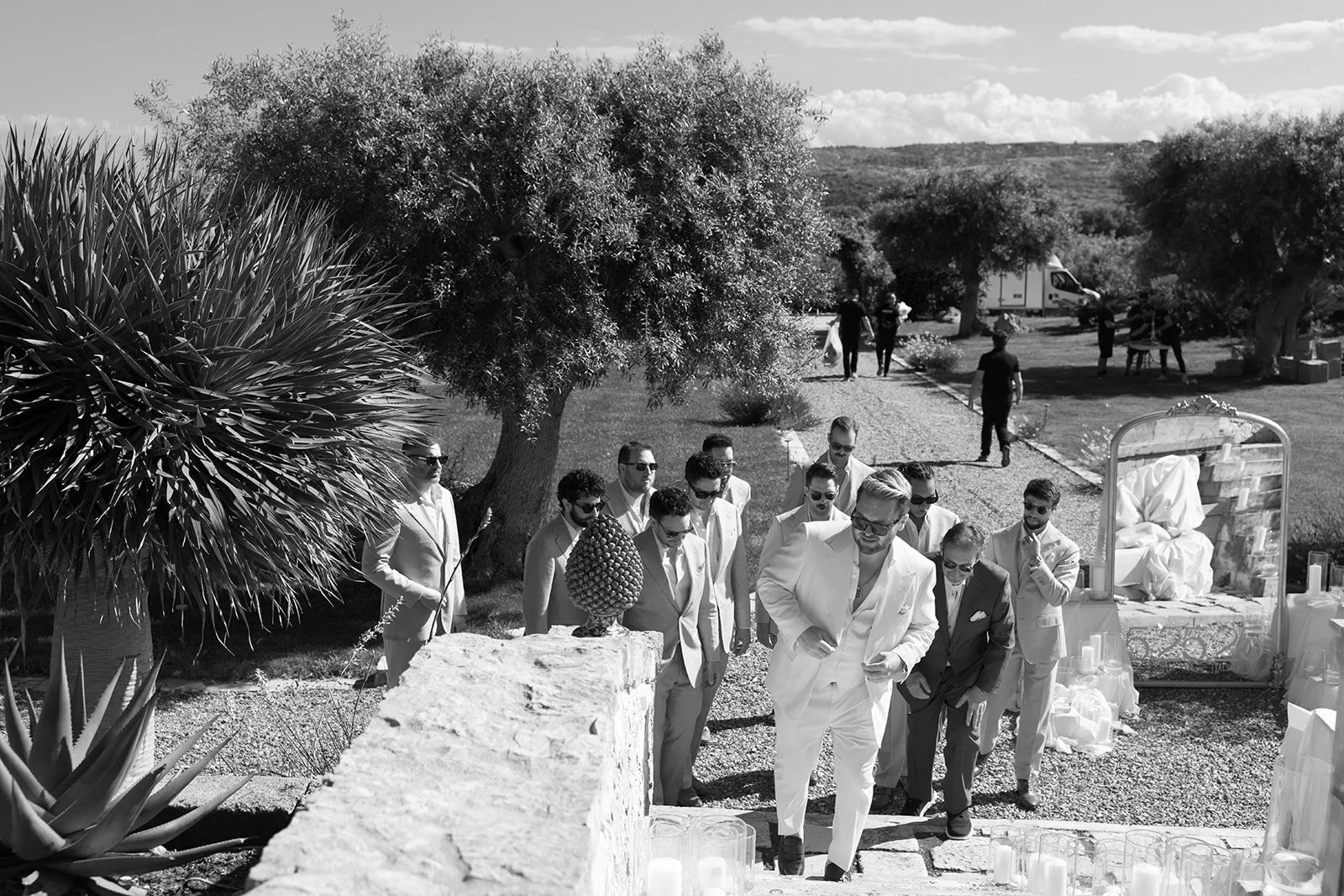 A group of men in light-colored suits, some with sunglasses, walking outdoors on a gravel path during a sunny day, surrounded by trees and hills.